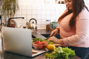 Overweight woman using laptop to watch video recipe while making vegan vitamin avocado salad, slicing leaf lettuce on wooden cutting board. Healthy food, weight loss, dieting and nutrition concept